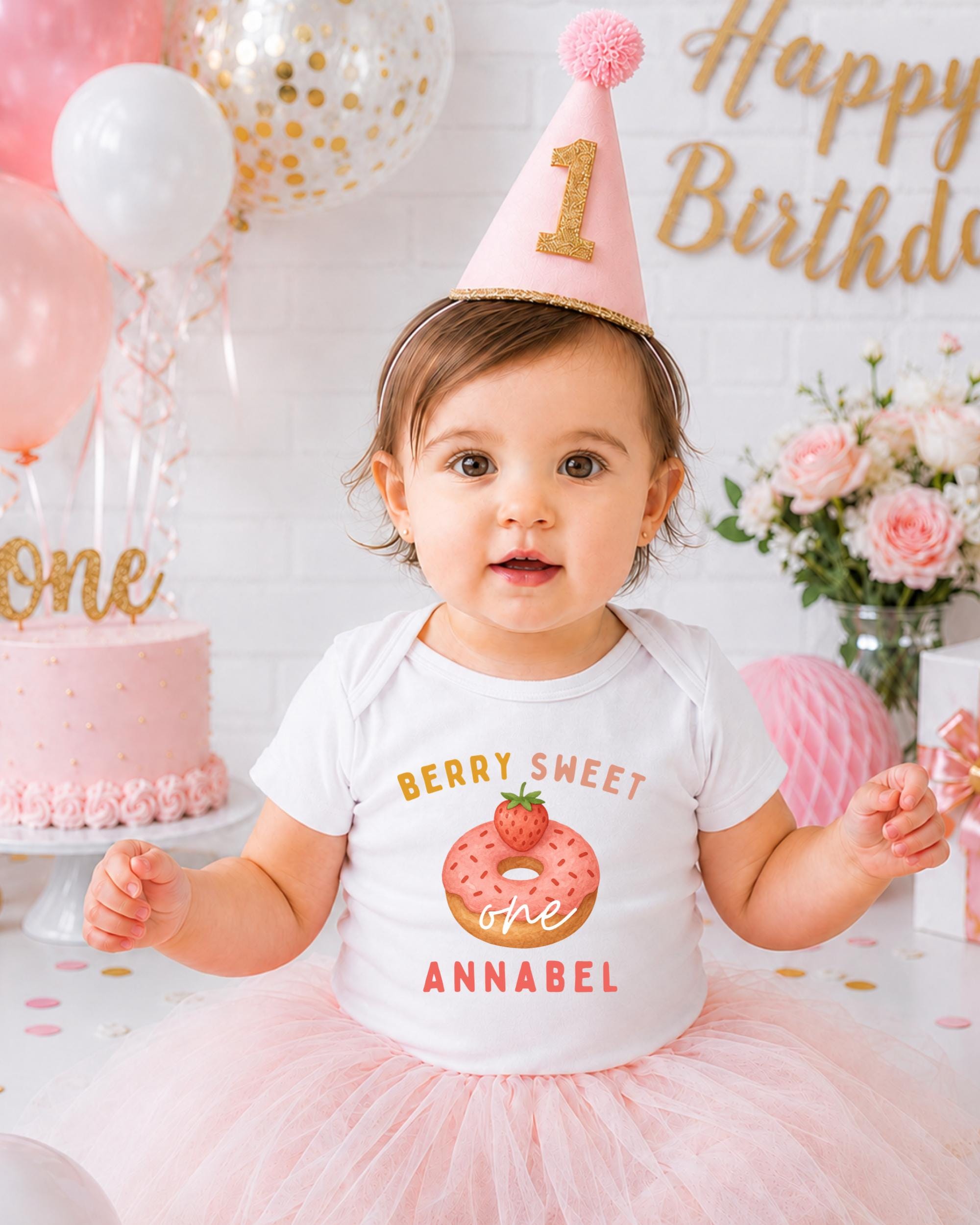 A young girl wearing a birthday hat and a white t-shirt with a strawberry donut graphic is posing in front of a birthday cake and balloons.