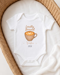 A white onesie with a coffee cup design and text printed on it, placed in a woven basket.