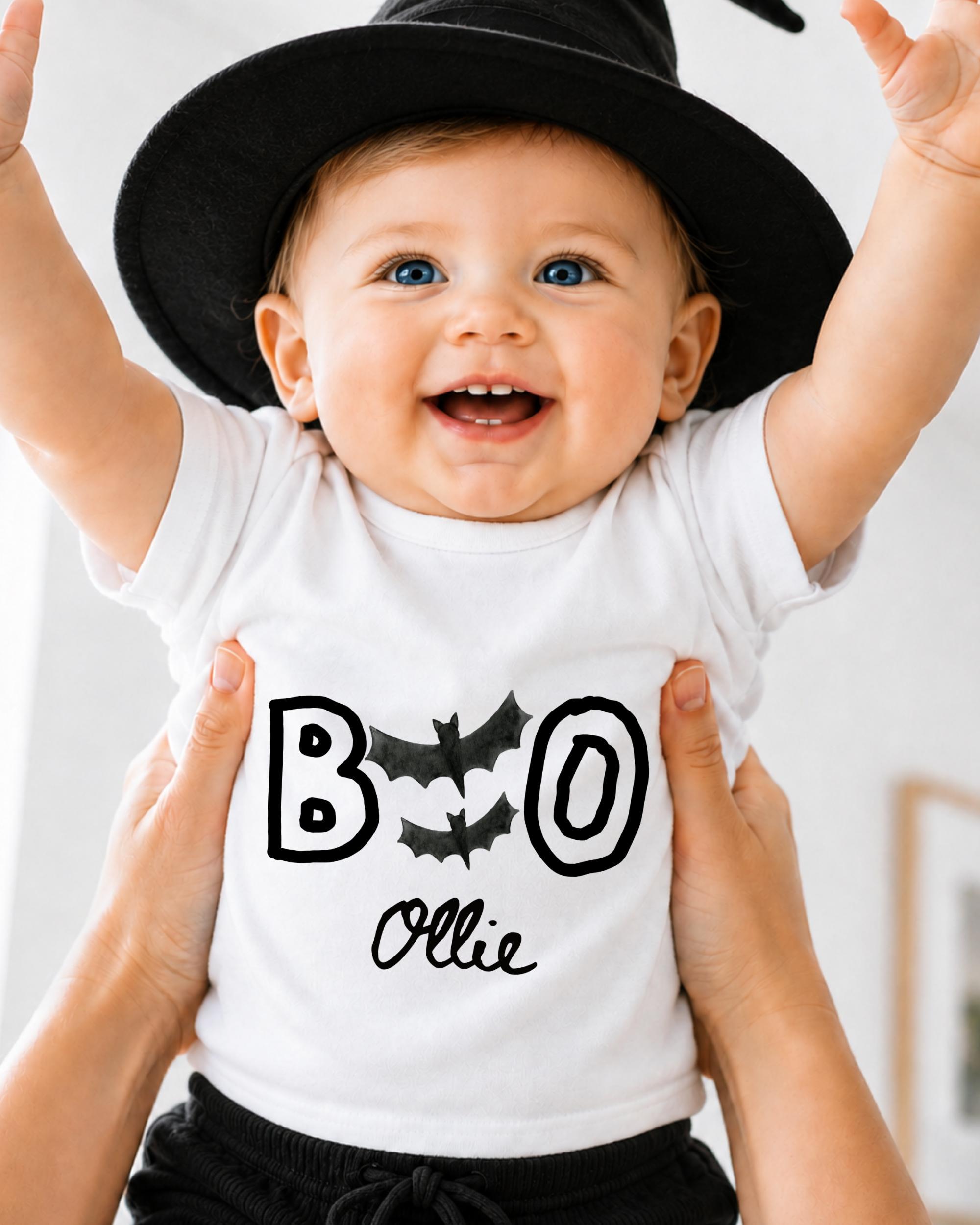 A smiling baby wearing a white t-shirt with the text "BOO OLLIE" and a black witch hat.