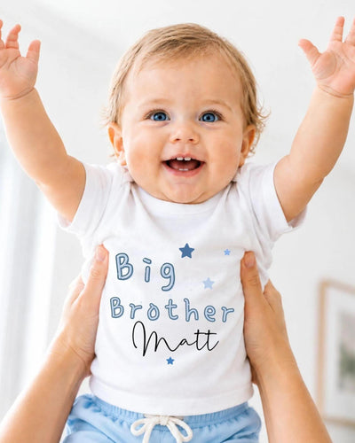 A smiling baby wearing a white shirt with the text "Big Brother Matt" and blue shorts, being held up by someone's arms.