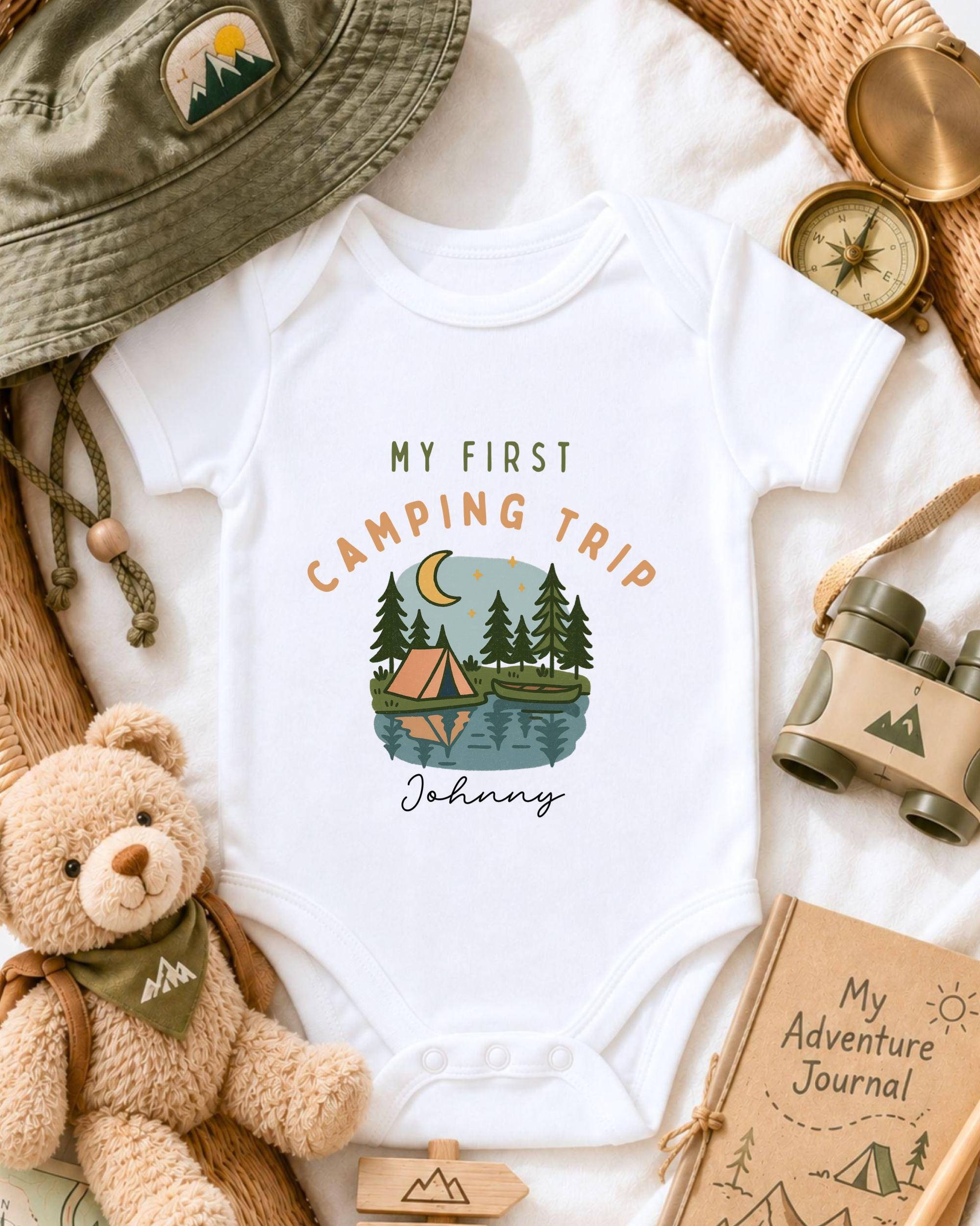 a baby onesie with a camping-themed design, surrounded by camping-related items such as a hat, compass, binoculars, and a teddy bear.