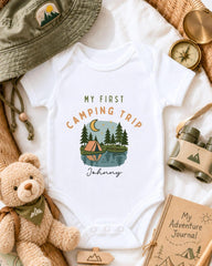 a baby onesie with a camping-themed design, surrounded by camping-related items such as a hat, compass, binoculars, and a teddy bear.