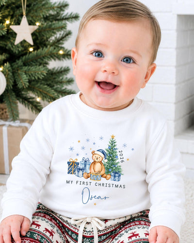 A smiling baby boy wearing a white sweater with a Christmas-themed design and text, sitting in front of a Christmas tree.