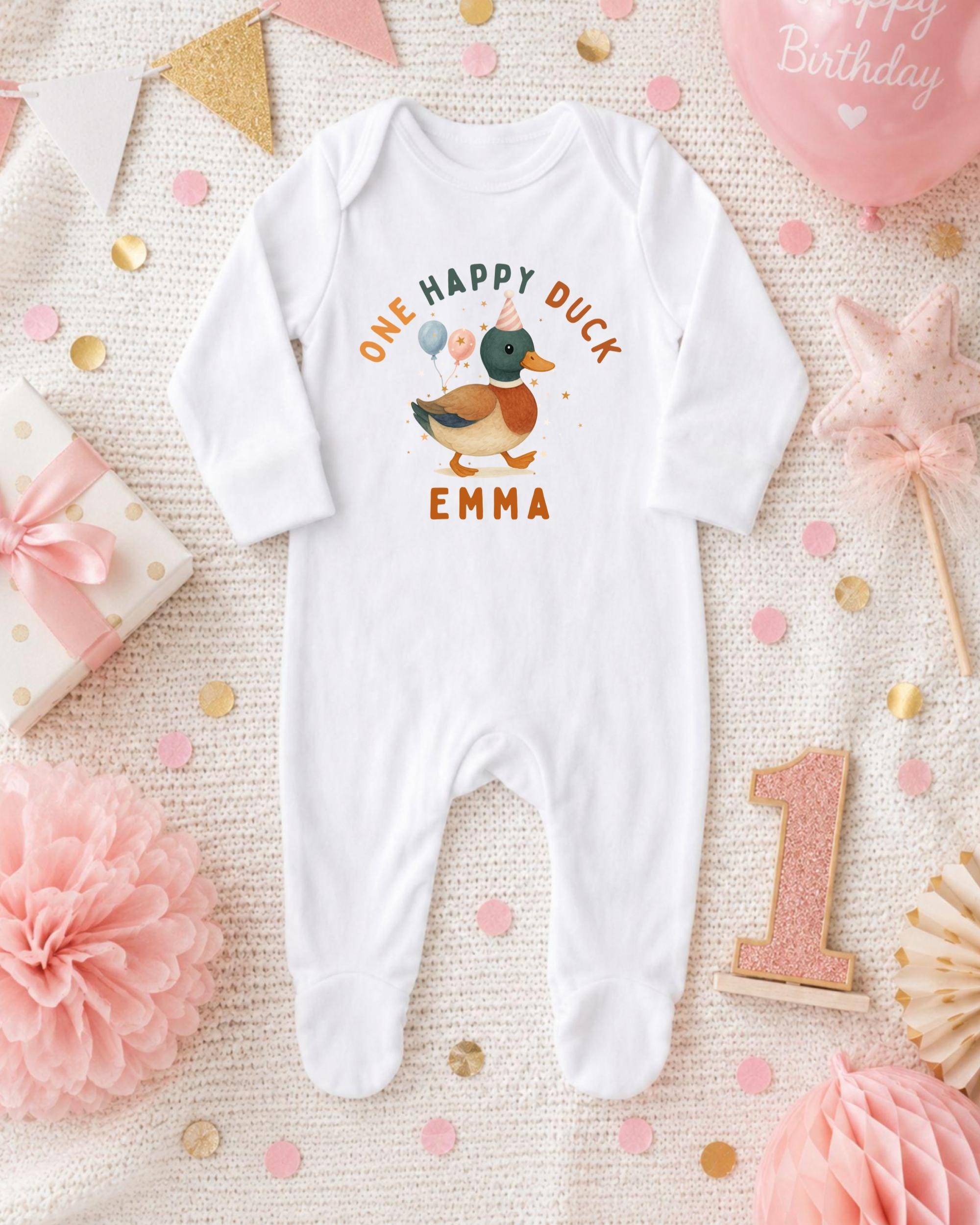 A white onesie with a duck design and the text "One happy duck Emma" printed on it, surrounded by birthday decorations and confetti.