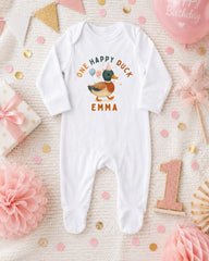 A white onesie with a duck design and the text "One happy duck Emma" printed on it, surrounded by birthday decorations and confetti.
