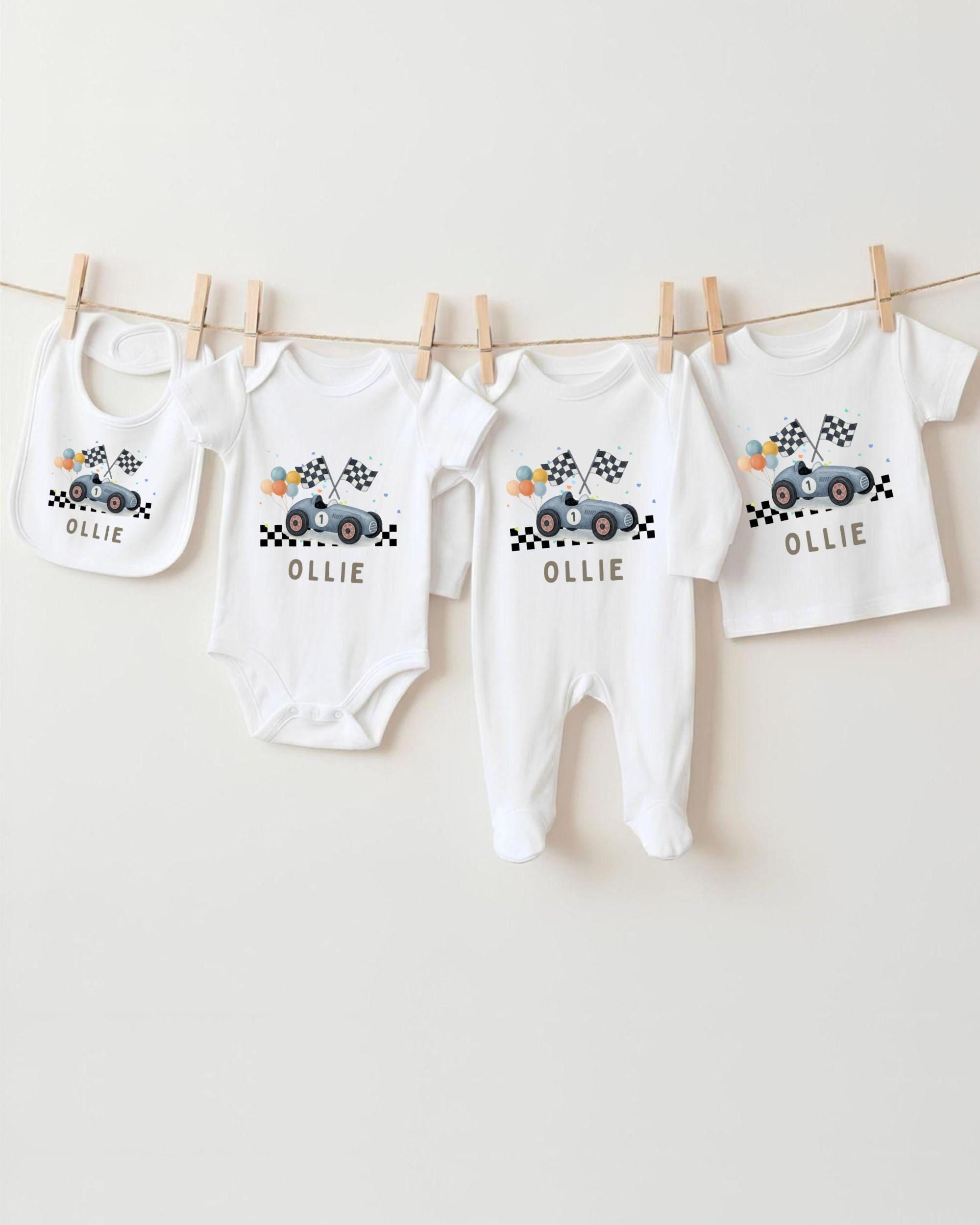 A collection of white baby clothes, including onesies and bibs, with a racing car design, hanging on a clothesline.