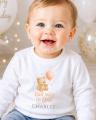 A smiling baby boy wearing a white shirt with a teddy bear and a balloon, sitting in front of birthday balloons.