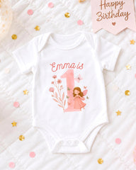 A white onesie with a pink fairy design and the number 1, surrounded by pink and gold confetti.