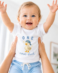A smiling baby wearing a white shirt with a cartoon duck and the text "ONE cool duck" is being held up by someone's arms.