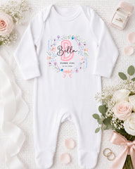 A white onesie with a floral design and the name "Bella" printed on it, along with a pink ribbon and a bouquet of white flowers.