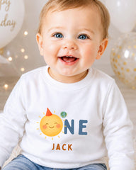 A smiling baby boy wearing a white shirt with a sun and planet graphic and the name "Jack" printed on it.