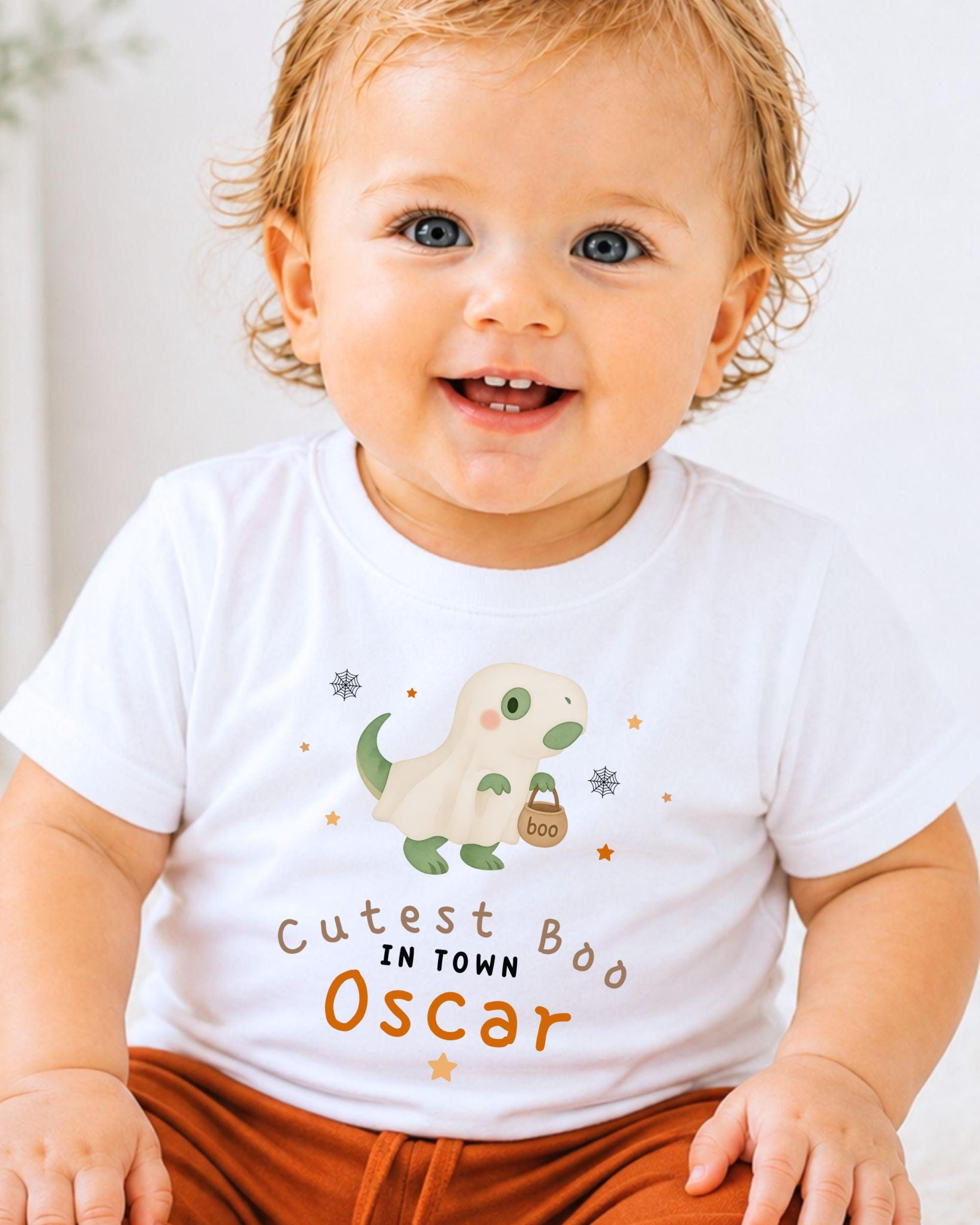 A smiling baby boy wearing a white t-shirt with a cartoon dinosaur and text that says "Cutest Boo Oscar".