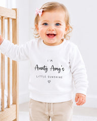A smiling baby girl wearing a white sweatshirt with text and a heart symbol.