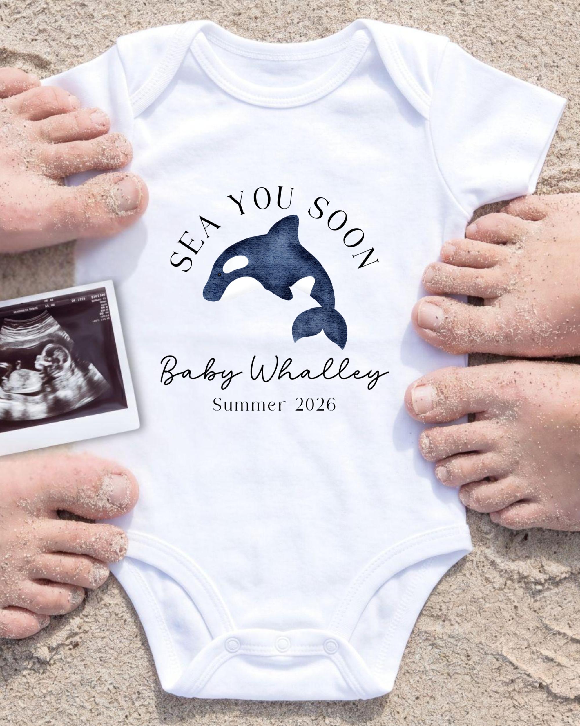 A white onesie with a graphic of a whale and the text "SEA YOU SOON Baby Whalley Summer 2026" is displayed on a sandy beach, surrounded by people's hands.