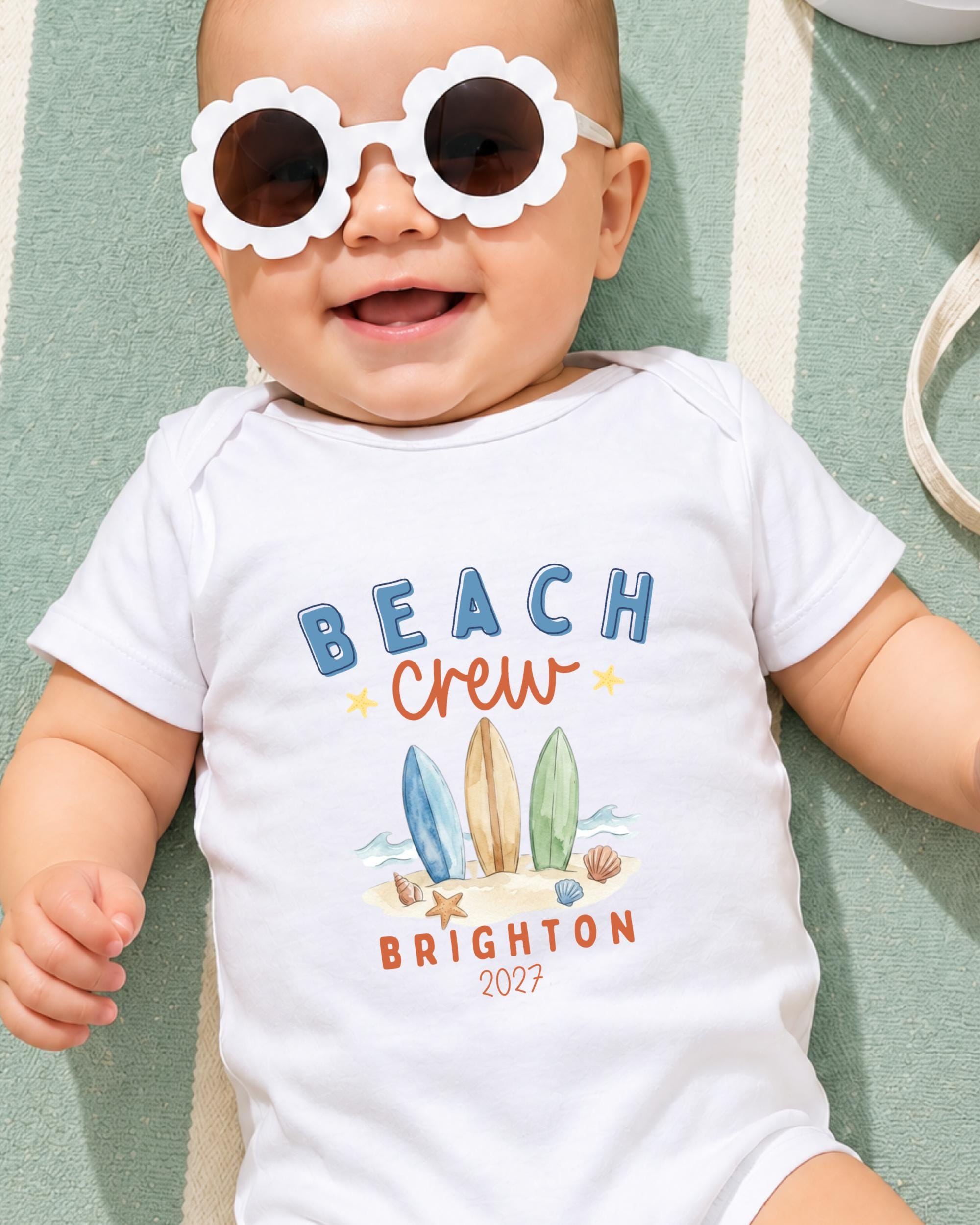 A smiling baby wearing a white onesie with a beach crew design and sunglasses, lying on a green and white striped surface.