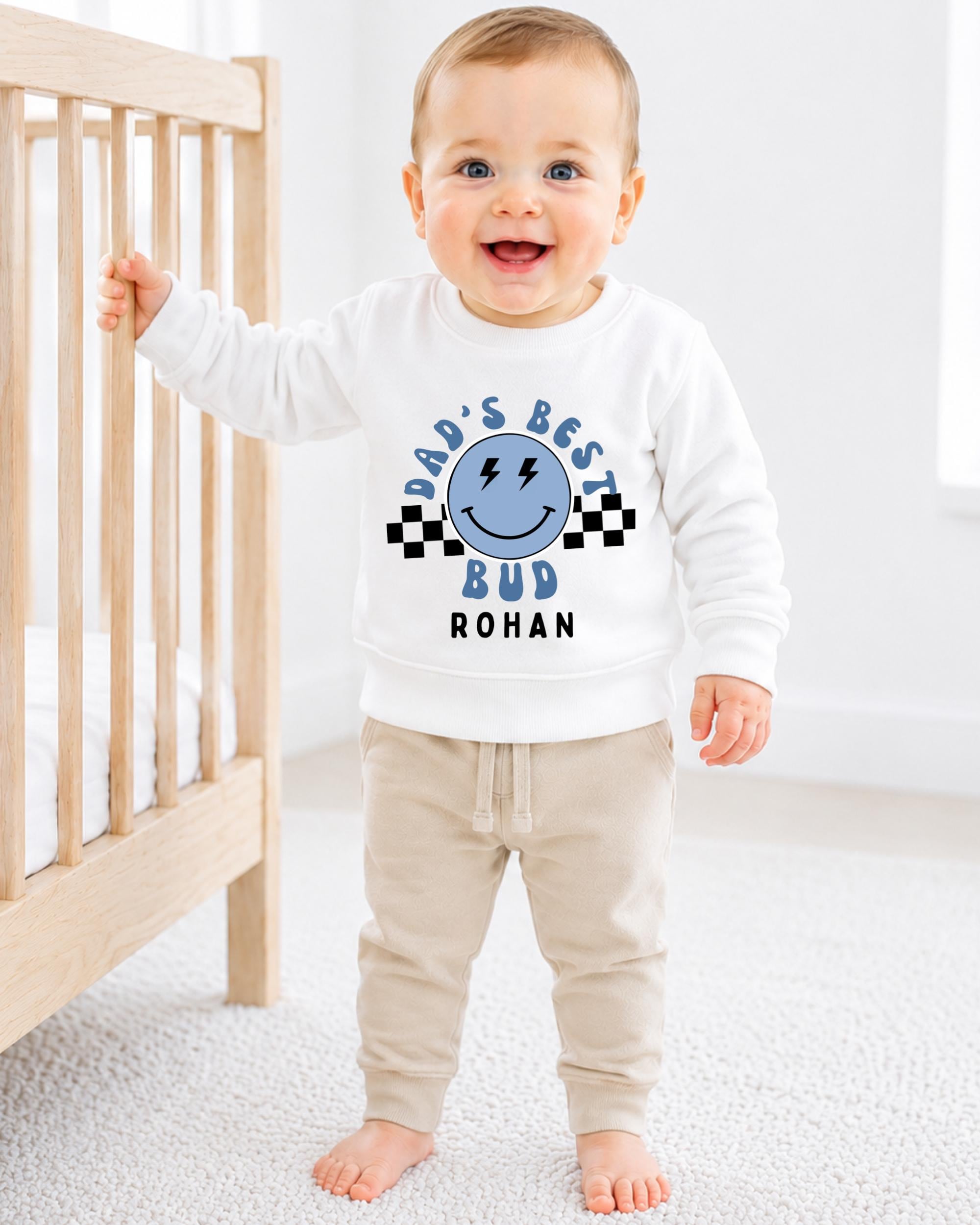 A smiling baby boy wearing a white sweatshirt with a graphic design and text, standing in front of a wooden crib.