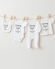 a row of four white baby clothes hanging on a clothesline, each with a black text that announces the upcoming arrival of a baby.