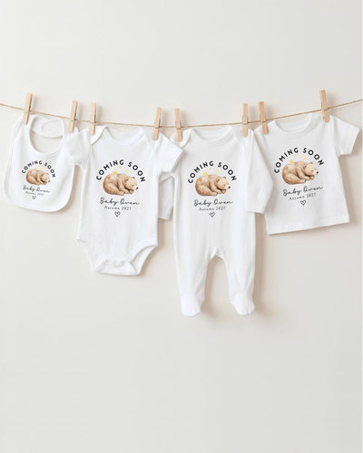 Personalisable white Bib, Baby Vest, Baby Grow and T Shirt with a sleeping brown watercolour bear beneath curved black 'Coming Soon' lettering, 'Baby Owen Autumn 2027' text and an outlined heart.