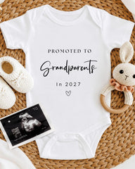A white baby onesie with the text "Promoted to Grandparents In 2027" printed on it, along with a crocheted rabbit toy and a black and white photograph of an Ultrasound image.