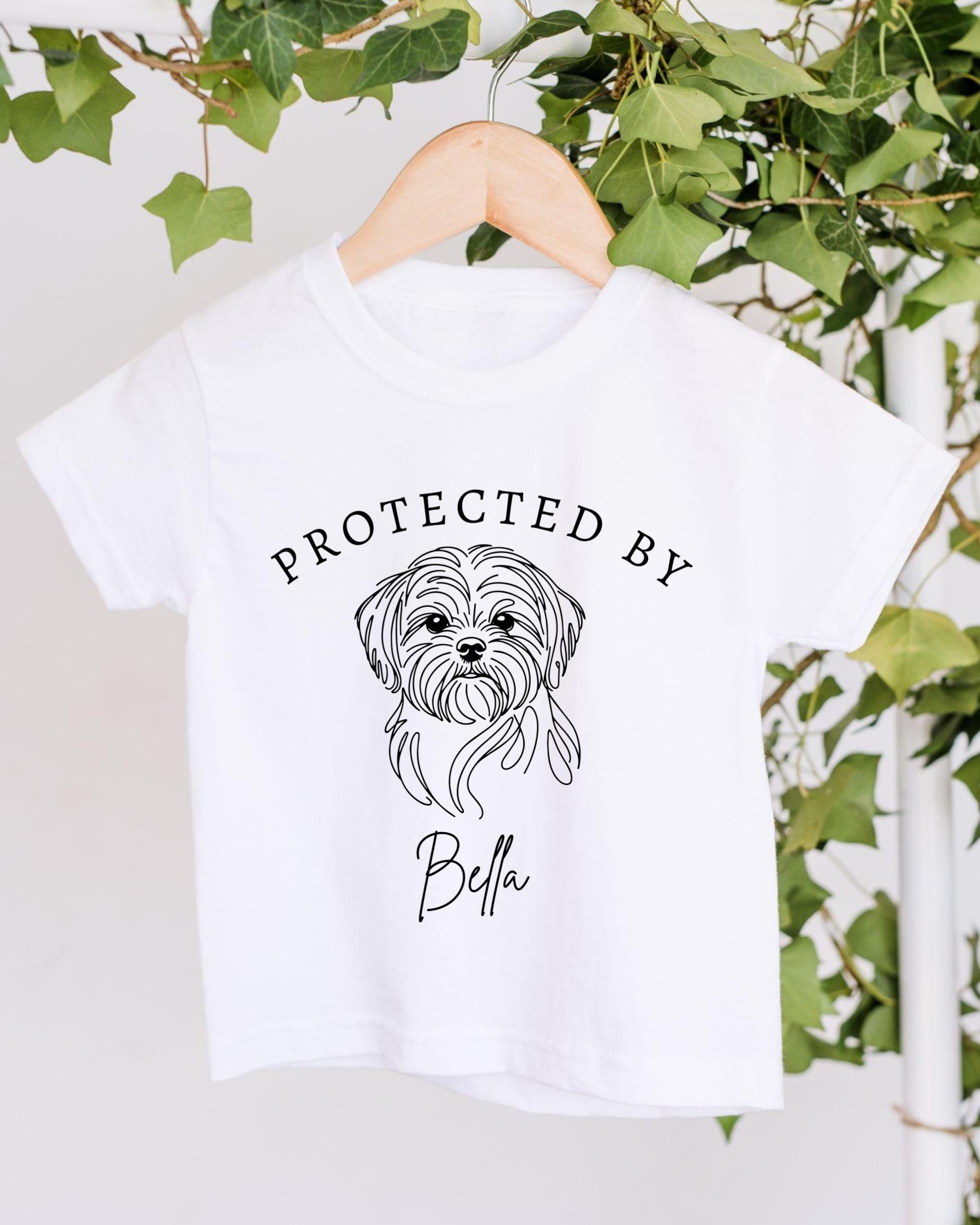 A white t-shirt with a black graphic of a dog's face and the text "PROTECTED BY Bella" printed on it.