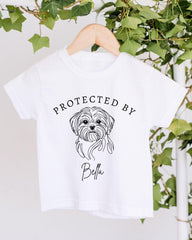 A white t-shirt with a black graphic of a dog's face and the text "PROTECTED BY Bella" printed on it.
