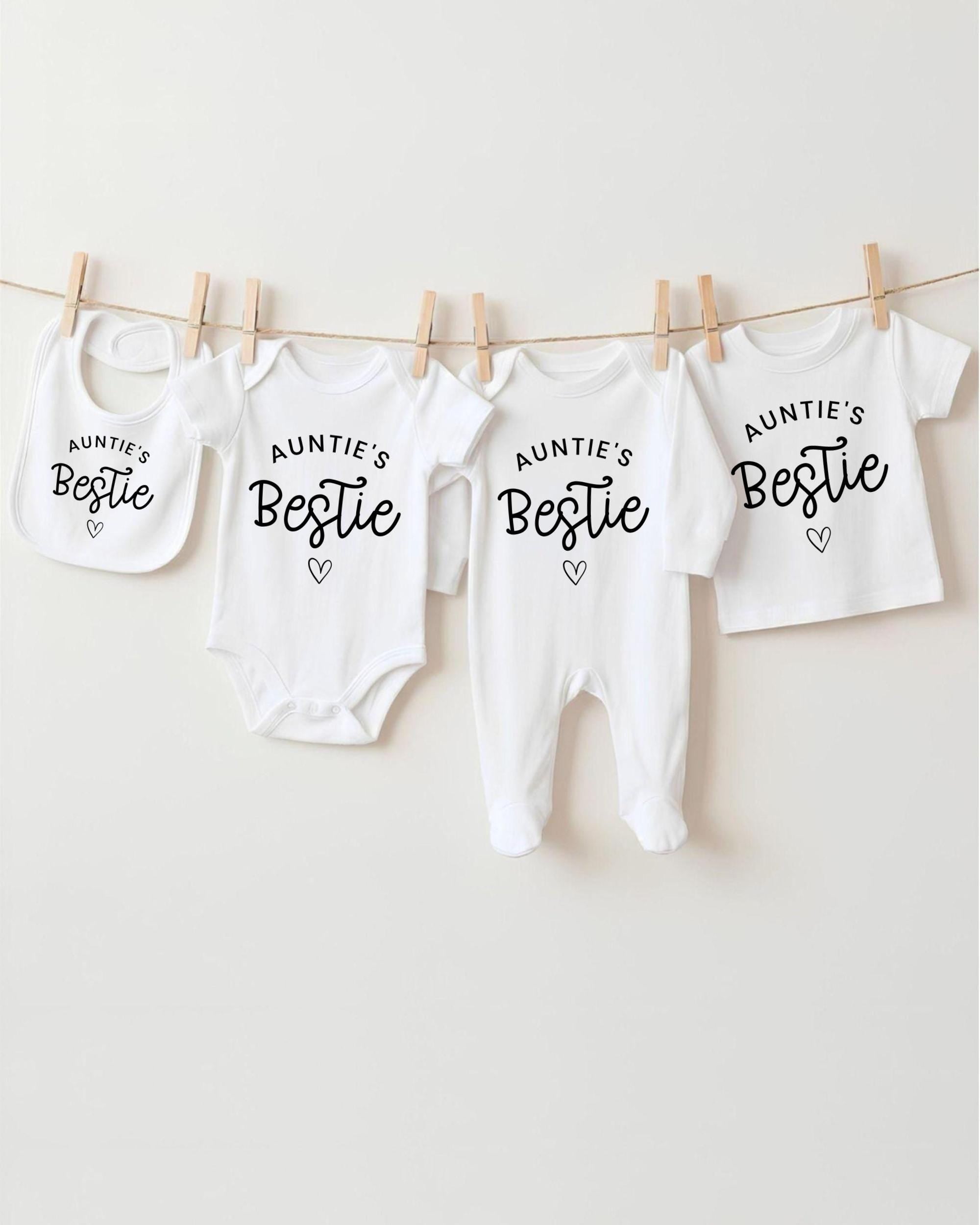 A row of white baby clothes with the text "Auntie's Bestie" printed on them, along with a bib and a pair of white onesies.