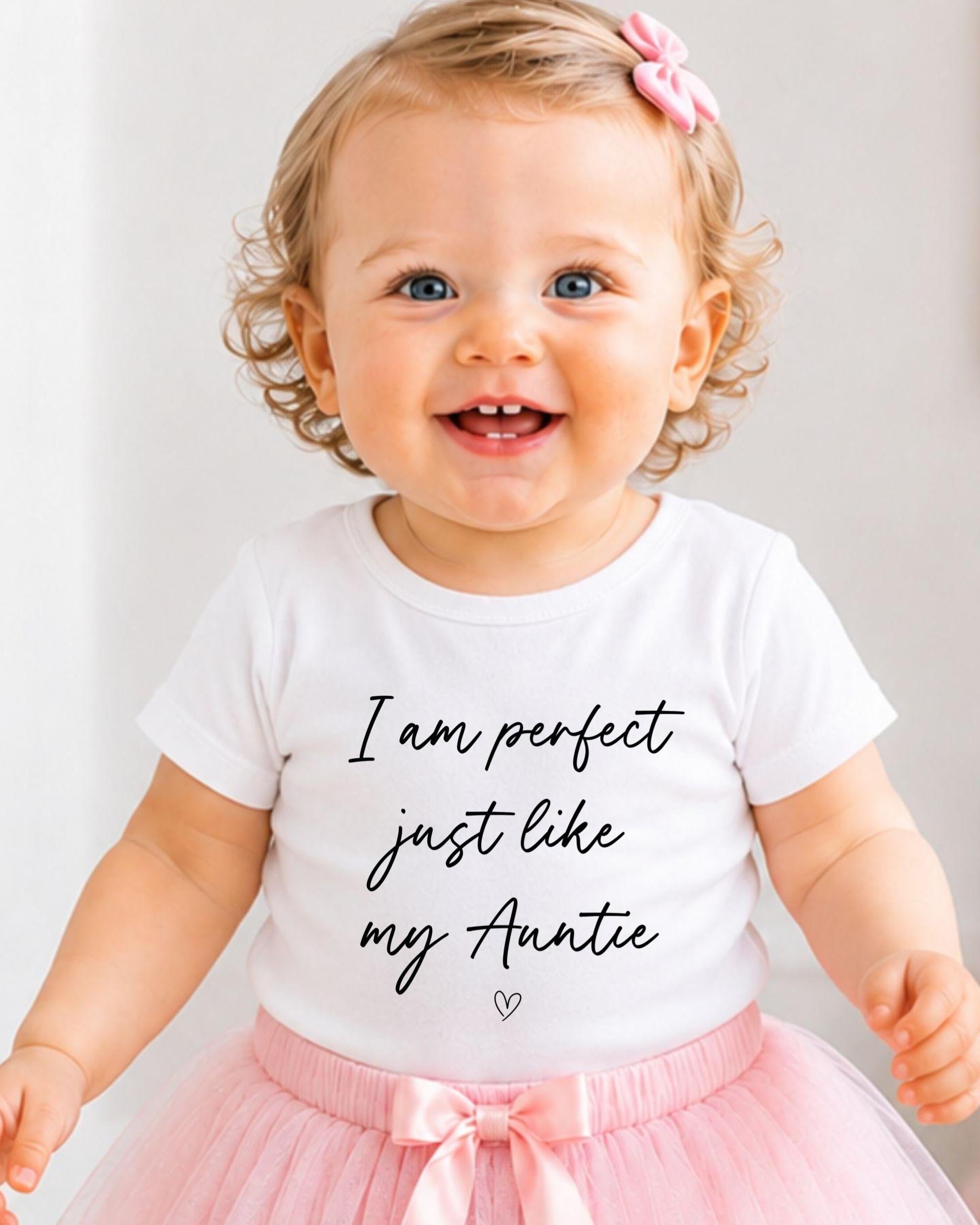 A smiling baby girl wearing a white t-shirt with text and a pink bow in her hair.