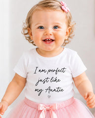 A smiling baby girl wearing a white t-shirt with text and a pink bow in her hair.