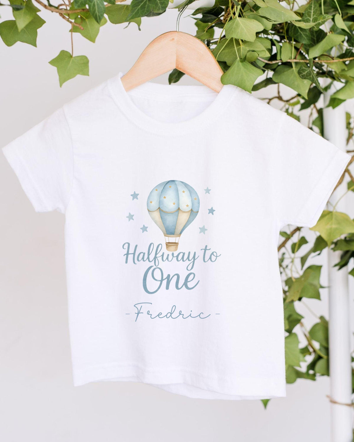 A white t-shirt with a printed design of a hot air balloon and stars, along with the text "Halfway to One - Frederic".