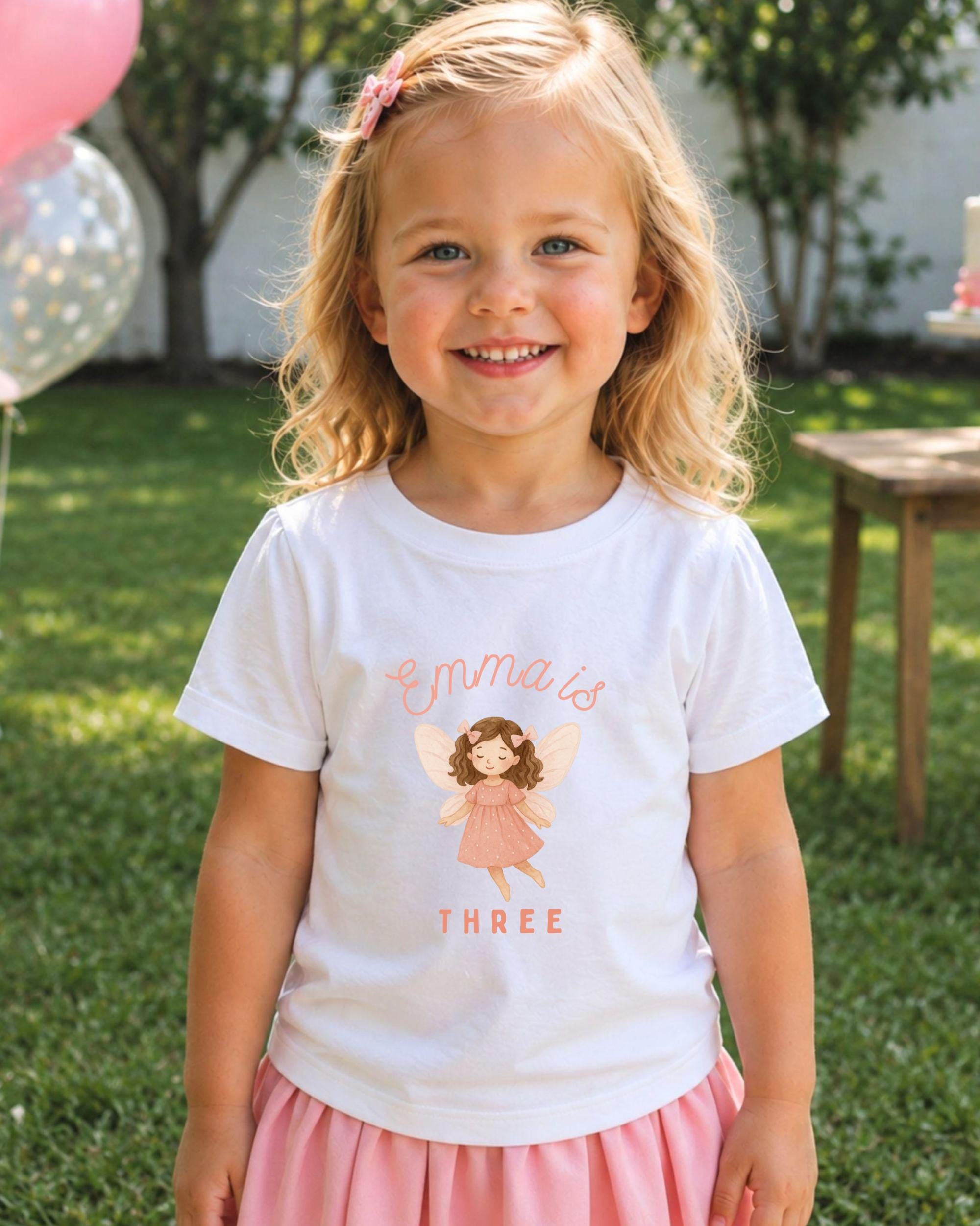 A young girl with blonde hair is smiling and wearing a white t-shirt with a fairy graphic and the text "Emma is three".