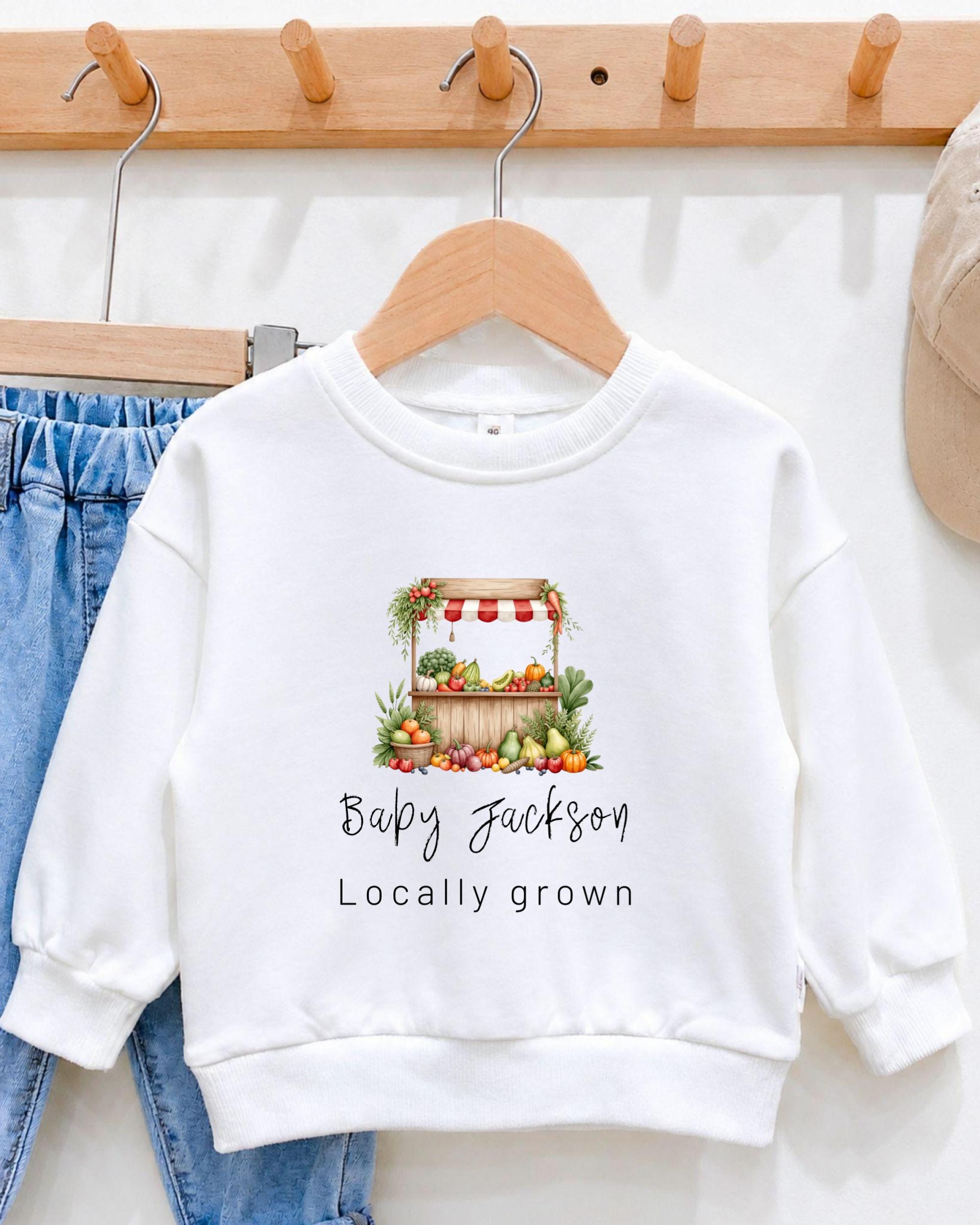 A white sweatshirt with a graphic of a fruit stand and the text "Baby Jackson Locally grown" printed on it, hanging on a wooden hanger.