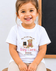 A young girl wearing a white t-shirt with a teddy bear and the text "My First Day of Daycare Annie September 2023" smiling at the camera.