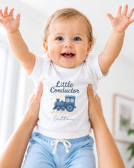A smiling baby wearing a white shirt with a train graphic and the name "Sutton" printed on it, being held up by someone's arms.