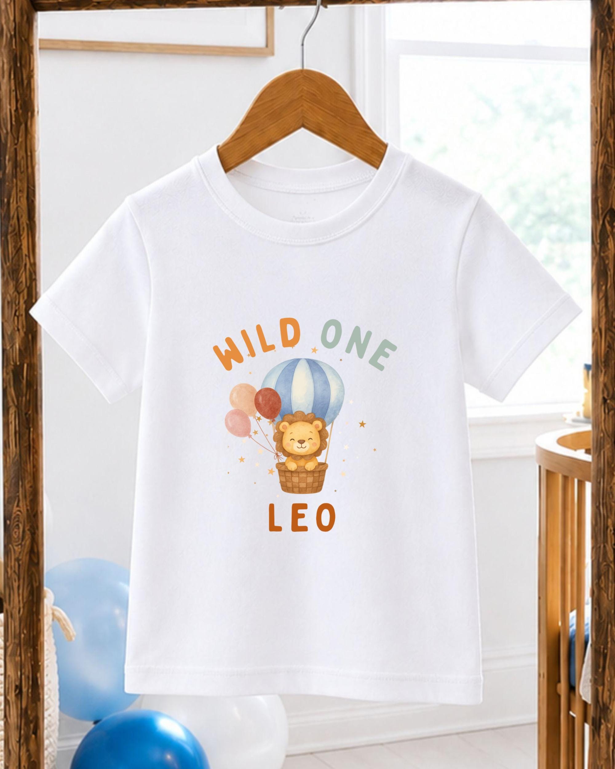 A white t-shirt with a printed design of a lion and balloons, along with the text "WILD ONE LEO".