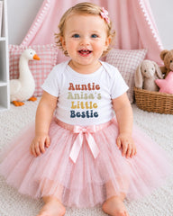A smiling baby girl wearing a white t-shirt with the text "Auntie Anisa's Little Bestie" and a pink tutu skirt, sitting on a white carpet surrounded by stuffed animals.