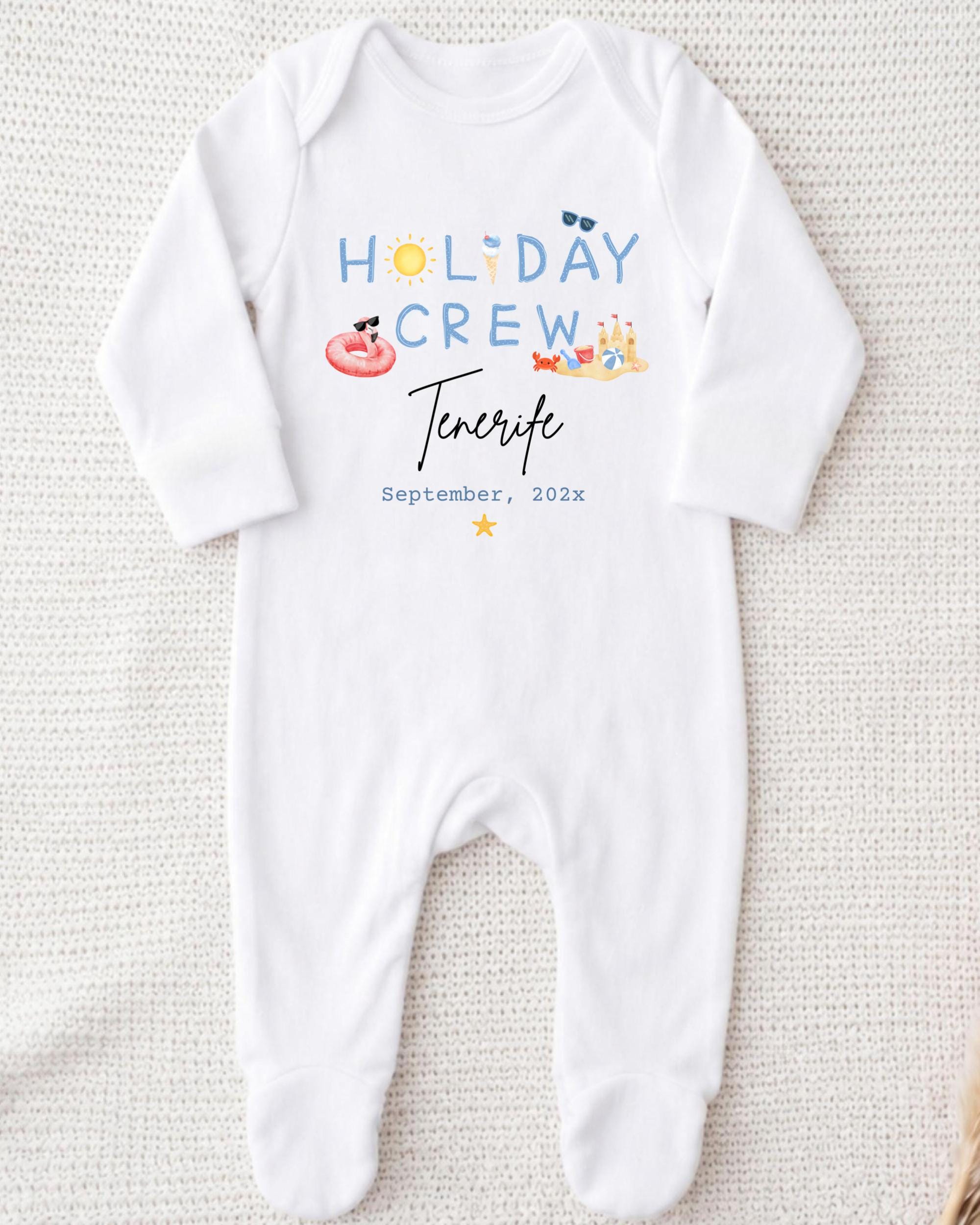 A white onesie with the text "HOLIDAY CREW" and "Tenerife" printed on it, along with illustrations of a sun, sunglasses, and a beach scene.