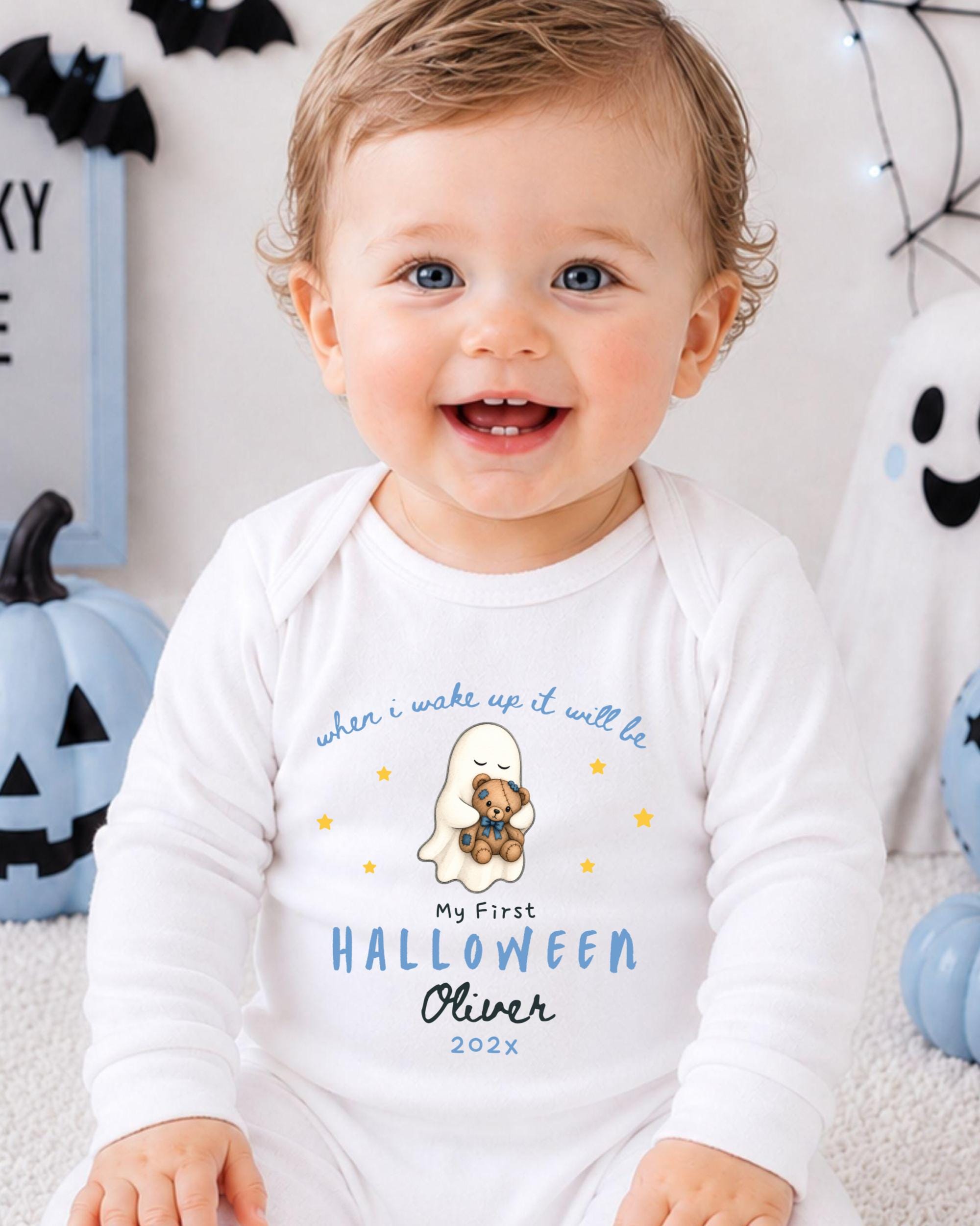 A smiling baby boy wearing a white onesie with a Halloween-themed design, sitting in front of Halloween decorations.