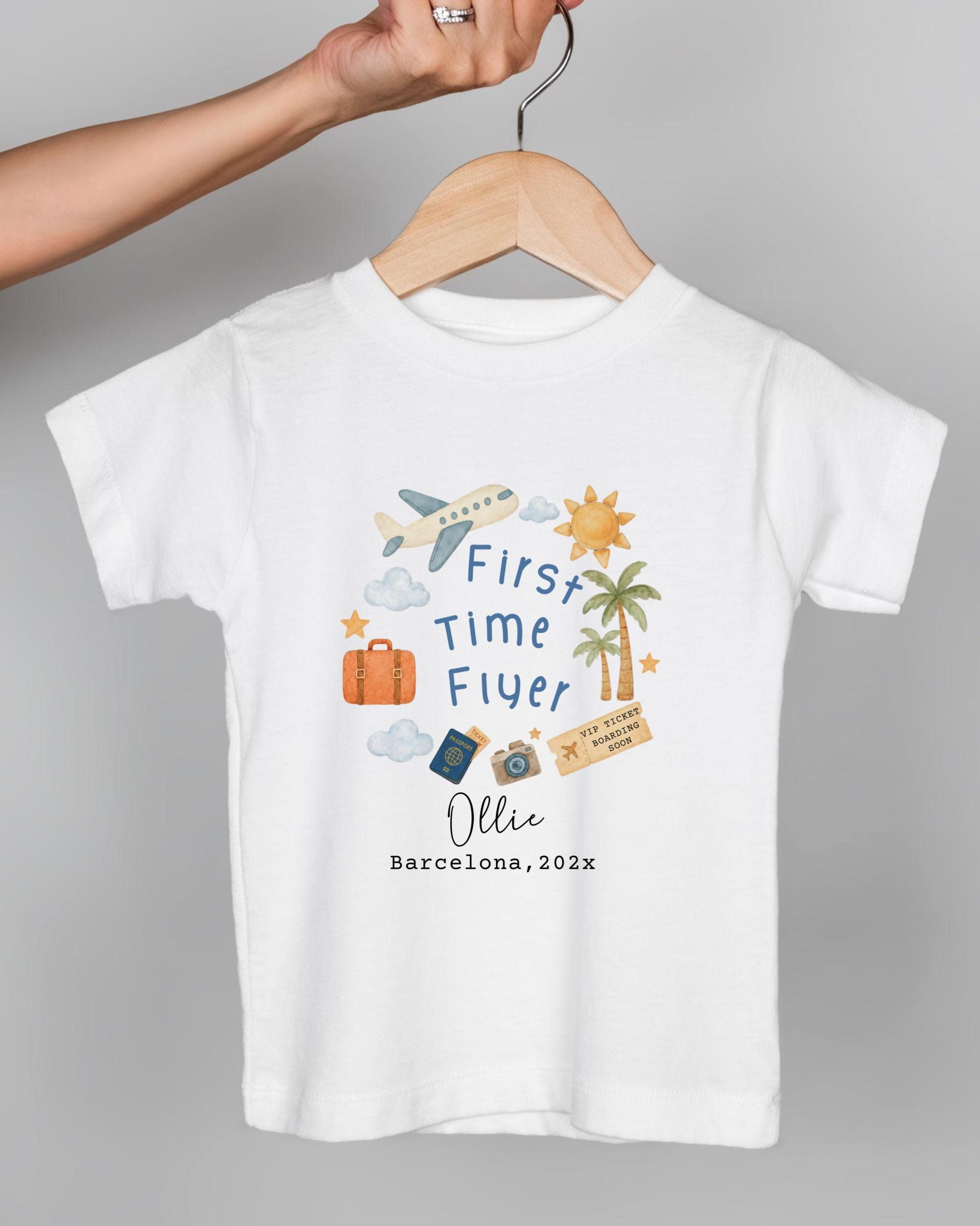 A white t-shirt with a graphic design featuring various travel-related icons, including a suitcase, a plane, a palm tree, and a sun. The text on the t-shirt reads "First Time Flyer Ollie Barcelona, 202x".