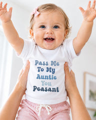 A smiling baby girl wearing a white t-shirt with text and a pink bow, being held up by someone's arms.