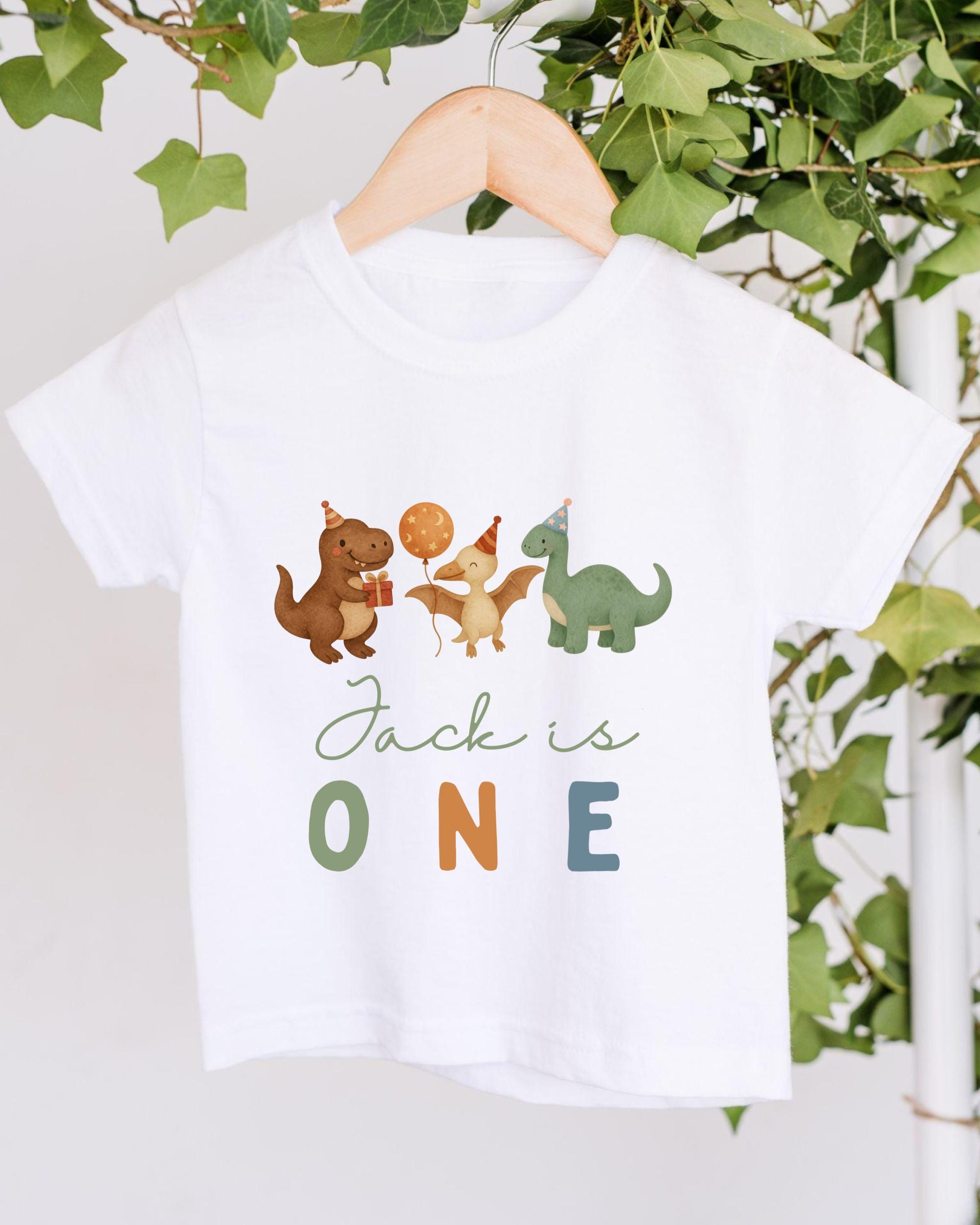 A white t-shirt with a graphic design featuring cartoon dinosaurs and the text "Jack is ONE" printed on it.