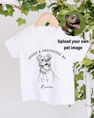 A white t-shirt with a black and white illustration of a dog and the text "LOVED & PROTECTED BY" printed on it.