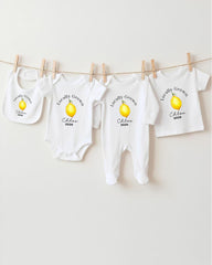 A row of white baby clothes with a yellow lemon graphic, each with a personalized name and year printed on them.