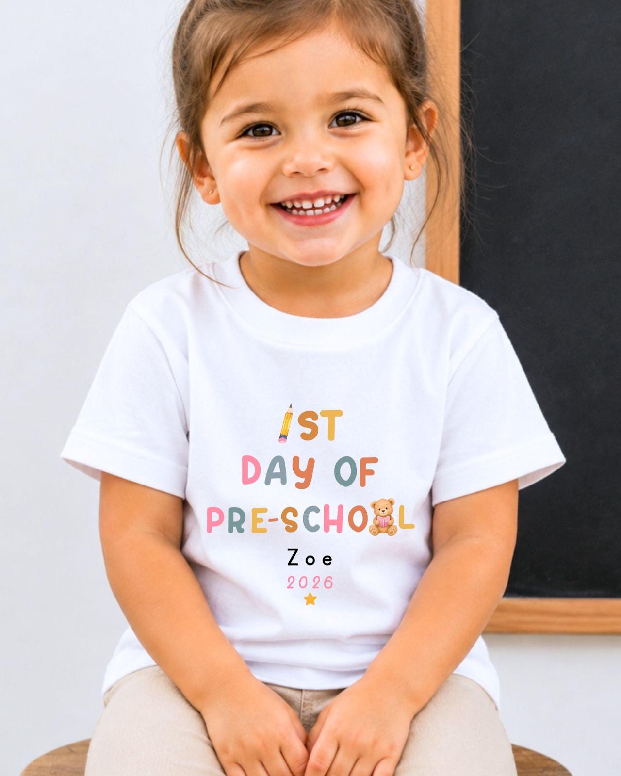 A young girl wearing a white t-shirt with text that says "1st DAY OF PRE-SCHOOL" and "Zoe 2026" printed on it, smiling at the camera.