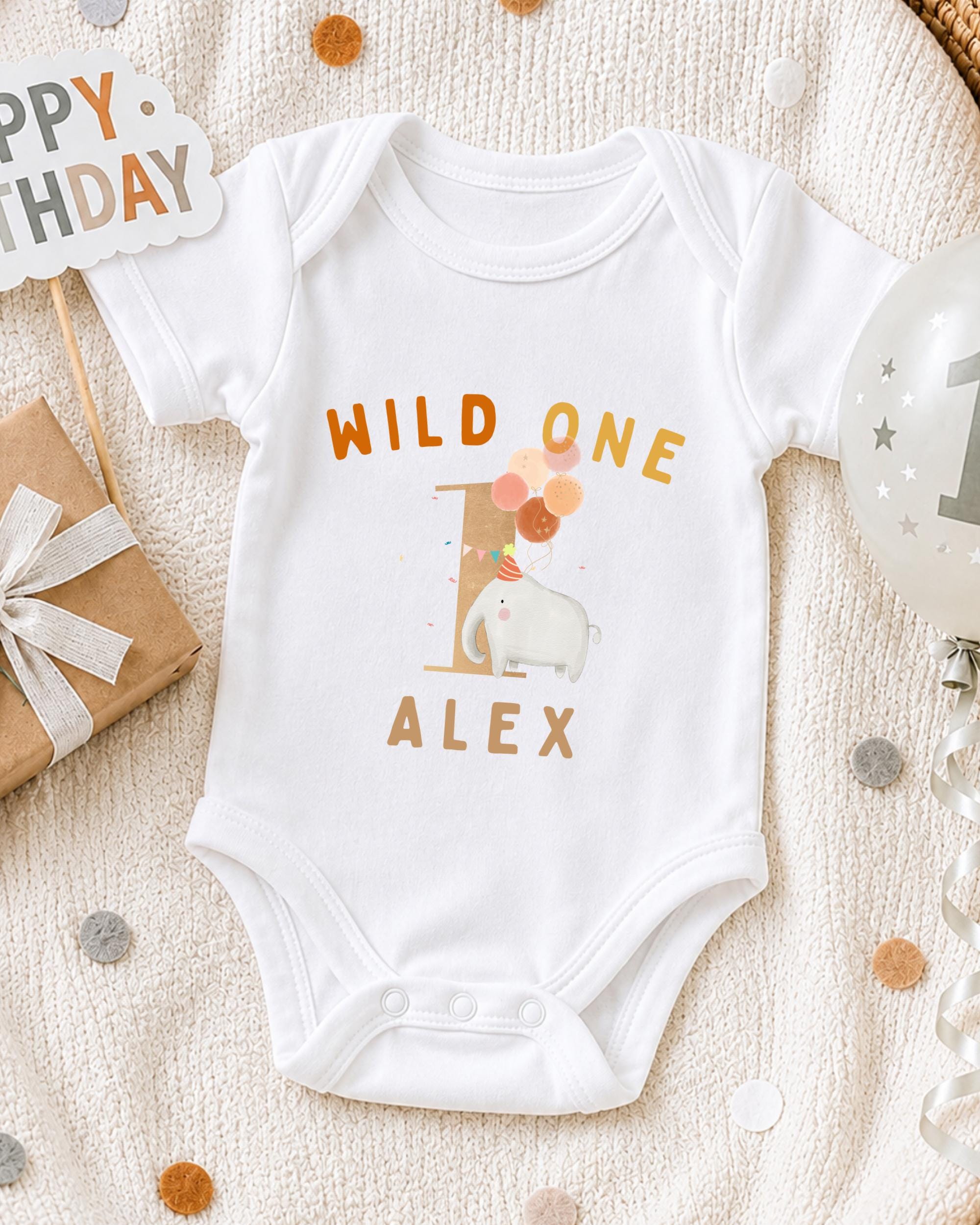 A white onesie with the text "WILD ONE ALEX" printed on it, along with an elephant and balloons, is displayed on a bed with a "Happy Birthday" sign and a birthday balloon.