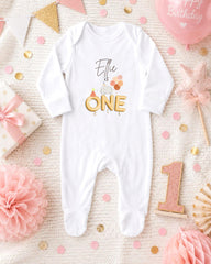A white onesie with the text "Elle is one" printed on it, surrounded by birthday decorations such as a pink star, confetti, and a pink balloon.