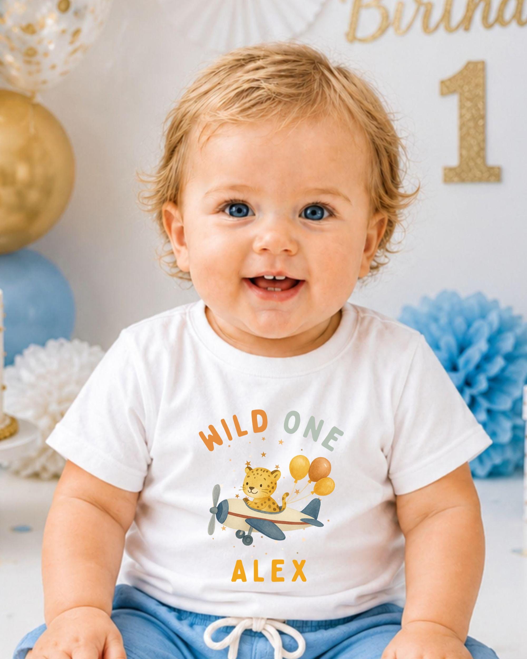 A smiling baby boy wearing a white t-shirt with a cartoon airplane and the text "Wild One Alex" is sitting in front of a birthday party backdrop.