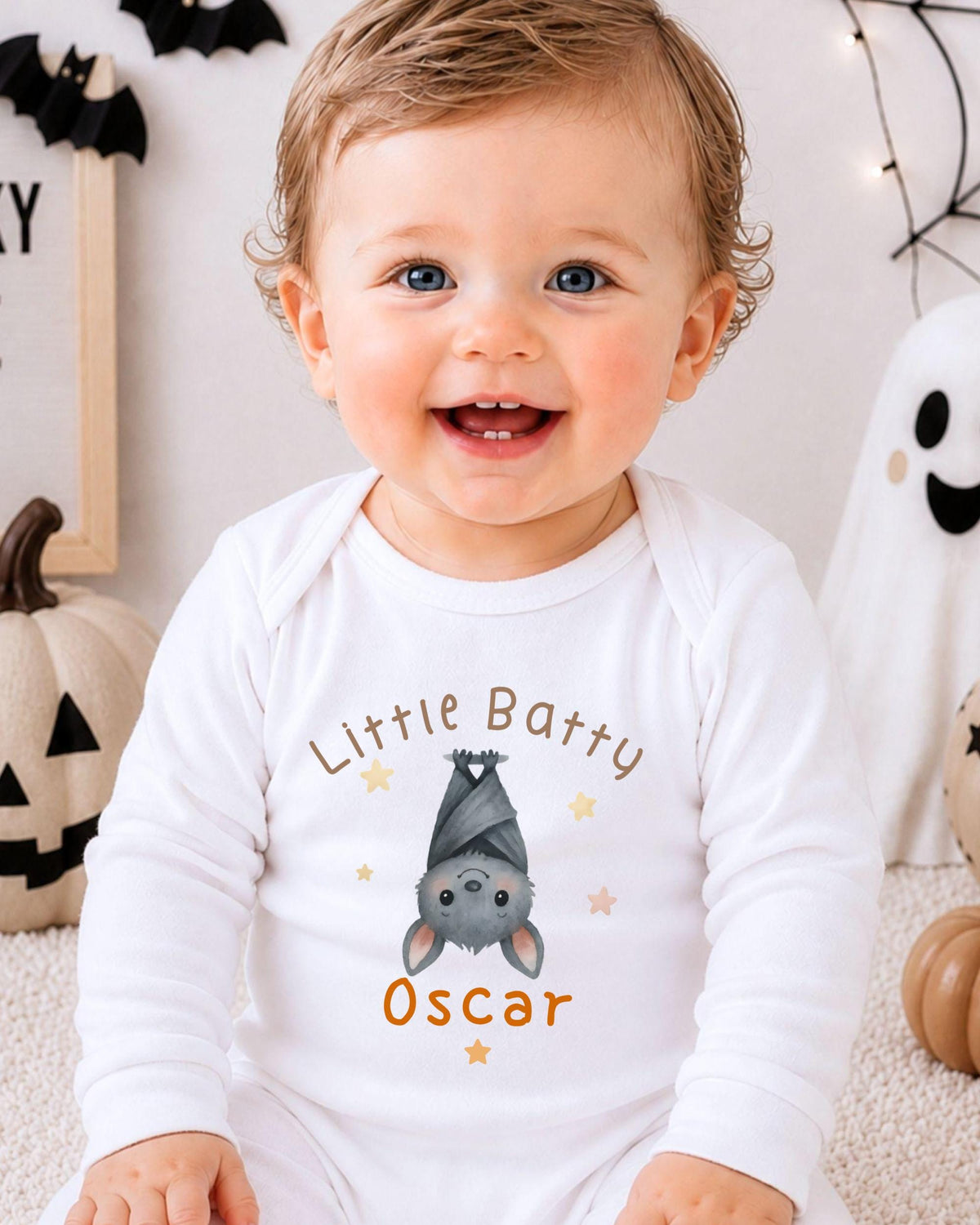 A smiling baby boy wearing a white onesie with a bat graphic and the name "Oscar" printed on it, sitting in front of Halloween decorations.