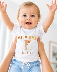 A smiling baby wearing a white t-shirt with the text "WILD ONE ALEX" and an elephant graphic, being held up by someone's arms.