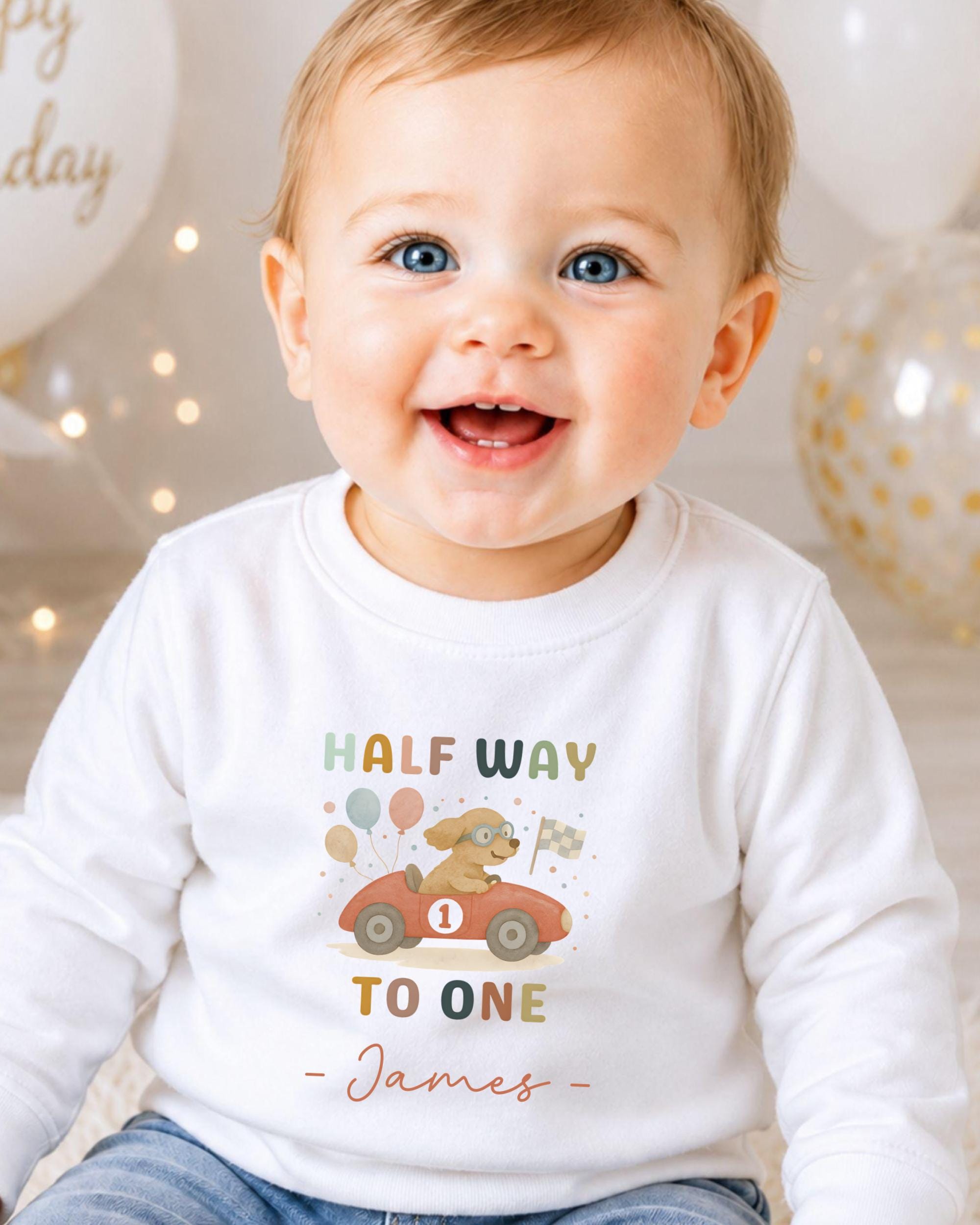A smiling baby boy wearing a white shirt with a colorful graphic and text, sitting in front of birthday balloons.