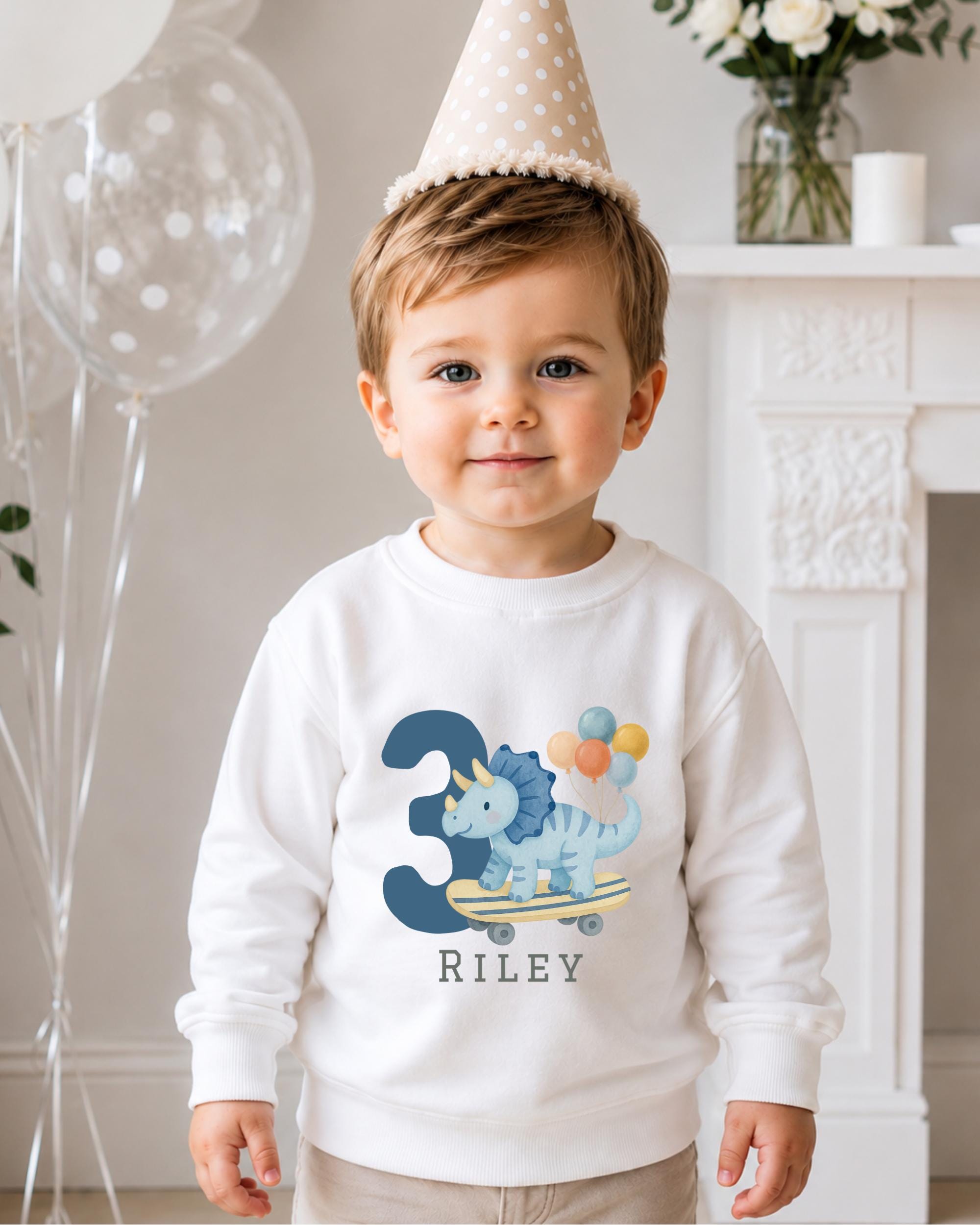 A young boy wearing a white sweatshirt with a dinosaur and balloons on it, standing in front of a fireplace with balloons and a vase of flowers.